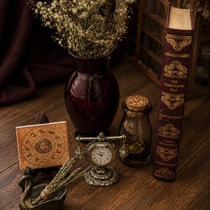 May include: A dark red glass vase filled with dried flowers, a square tile featuring a zodiac wheel, a small brass-toned desk clock, a glass jar with a cork stopper, and a hardcover book titled The Thirteenth Tale by Diane Setterfield.