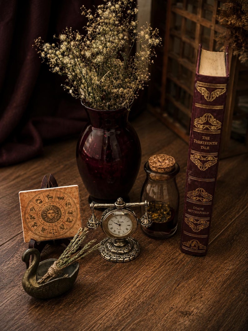 May include: A dark red glass vase filled with dried flowers, a square tile featuring a zodiac wheel, a small brass-toned desk clock, a glass jar with a cork stopper, and a hardcover book titled The Thirteenth Tale by Diane Setterfield.