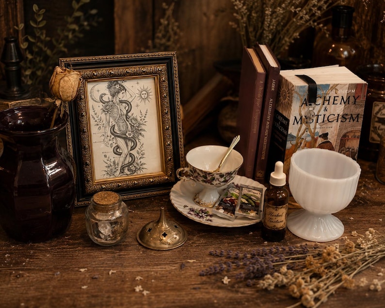 May include: A framed black and white illustration of a skeletal figure entwined with a snake, displayed alongside a dark purple vase, a teacup on a saucer, a book titled Alchemy & Mysticism, a small glass jar, and a white milk glass pedestal bowl.