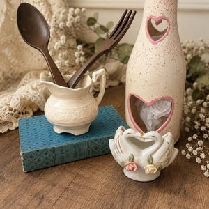 May include: A cream-colored ceramic pitcher containing a dark brown wooden spoon and fork, a tall speckled cream and pink bottle with heart-shaped cutouts, and a small white ceramic swan figurine with pink and yellow rose accents.