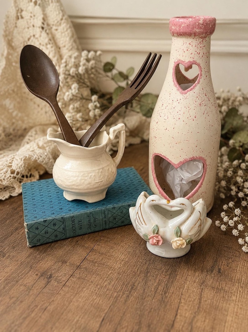 May include: A cream-colored ceramic pitcher containing a dark brown wooden spoon and fork, a tall speckled cream and pink bottle with heart-shaped cutouts, and a small white ceramic swan figurine with pink and yellow rose accents.