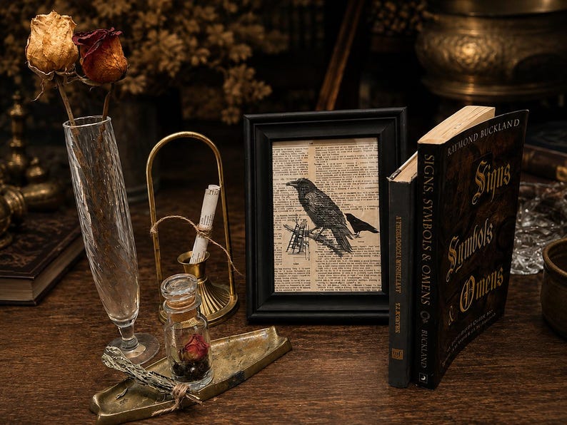 May include: A black picture frame containing a print of a crow on a dictionary page, positioned next to a book titled Signs, Symbols & Omens by Raymond Buckland, a tall textured glass vase with two dried roses, and a small brass tray with a glass vial.