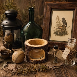 May include: A collection of items including a black metal lantern, a dark green glass bottle, a wooden picture frame with a raven and fern print, a bark-covered candle holder, a small metal cup, a smooth stone, and two small glass apothecary jars.