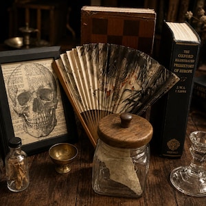 May include: A black framed print of a human skull over dictionary text, a folding fan with black ink patterns, a glass jar with a wooden lid, a small brass bowl, a clear glass bottle with contents, and a glass candlestick holder.
