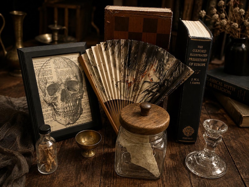 May include: A black framed print of a human skull over dictionary text, a folding fan with black ink patterns, a glass jar with a wooden lid, a small brass bowl, a clear glass bottle with contents, and a glass candlestick holder.