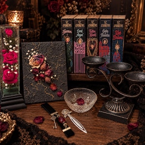 May include: A collection of items including a glass display case with three pink roses, a black textured canvas with dried flowers, five V.C. Andrews books, a metal triple candle holder, a clear glass bowl, a black bookmark with a key, and a metal rose bookmark.