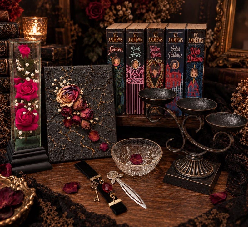 May include: A collection of items including a glass display case with three pink roses, a black textured canvas with dried flowers, five V.C. Andrews books, a metal triple candle holder, a clear glass bowl, a black bookmark with a key, and a metal rose bookmark.