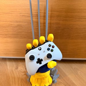 May include: A white Xbox wireless controller held by a yellow and blue hand-shaped stand featuring three long grey claws extending upwards. The base of the stand is yellow with brown accents and includes grey jagged rock-like elements at the bottom.