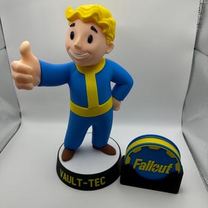 May include: A Vault Boy figurine with yellow hair, wearing a blue and yellow jumpsuit, standing on a circular base labelled VAULT-TEC. Beside it is a black holder containing several blue and yellow circular coasters with the text Fallout.