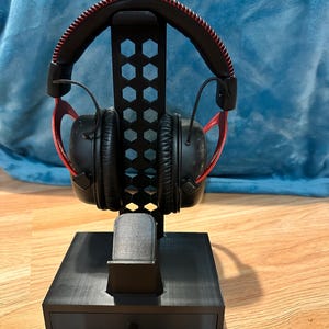 May include: Black gaming headphones with red detailing sit on a black headphone stand. The stand features a hexagonal design and a small drawer. The headphones have a black headband with red stitching and black earcups.