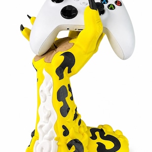 May include: A white wireless game controller held by a yellow, black, and white claw-shaped stand. The stand features a textured, organic design with black spots and a white flame-like pattern rising from the base to the fingers with black tips.