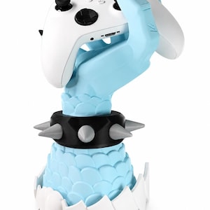May include: A light blue sculpted hand with black claws holding a white wireless game controller. The base features a black spiked collar and a white scalloped edge. The controller has black analogue sticks, a black D-pad, and white buttons.