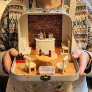 May include: A miniature bar scene inside a vintage suitcase. The interior features a brick wall, a white bar with bottles, a round table with chairs, and a small toolbox. The suitcase is held open, showcasing the detailed diorama.