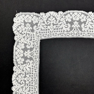 May include: Close-up of white lace trim with a floral pattern and scalloped edge. The lace is set against a black background, highlighting the intricate details of the design. Ideal for sewing and craft projects.