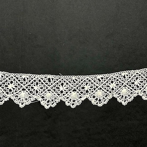 May include: A white lace trim featuring an intricate diamond-shaped grid pattern with small floral motifs centered within each diamond. The bottom edge of the trim is finished with a scalloped border, creating a decorative and delicate textile strip.