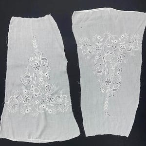 May include: Two white fabric pieces with intricate white floral embroidery. The embroidery features detailed flowers, leaves, and dotted accents. The edges of the fabric are unfinished, giving a vintage or handmade appearance. The pieces are set against a dark background.