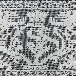 May include: Close-up of a white lace textile with a detailed, symmetrical design. The pattern features stylized floral and geometric elements, with a delicate, open weave. The edges are finished with a scalloped border.