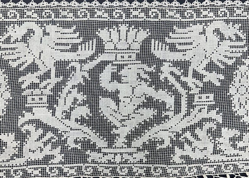 May include: Close-up of a white lace textile with a detailed, symmetrical design. The pattern features stylized floral and geometric elements, with a delicate, open weave. The edges are finished with a scalloped border.
