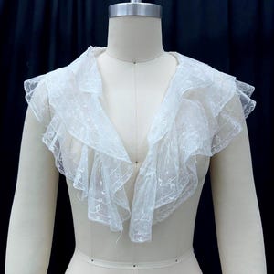 May include: A white, lacy collar with ruffled edges is displayed on a mannequin. The collar has a V-neck design and is made of a delicate, sheer fabric. The text "TSC" is visible at the bottom.