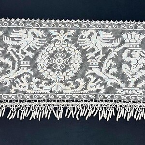 May include: A long, white lace runner with a detailed pattern of heraldic figures and floral motifs. The lace features a scalloped edge and a fringe along the bottom. The design includes eagles, crowns, and other ornate elements.