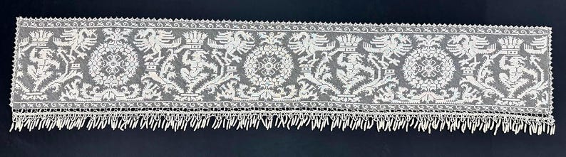 May include: A long, white lace runner with a detailed pattern of heraldic figures and floral motifs. The lace features a scalloped edge and a fringe along the bottom. The design includes eagles, crowns, and other ornate elements.