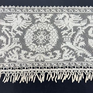 May include: Close-up of a white lace trim with a detailed floral and heraldic design. The lace features a repeating pattern of stylized crowns, floral motifs, and a central medallion. The bottom edge has a fringe.