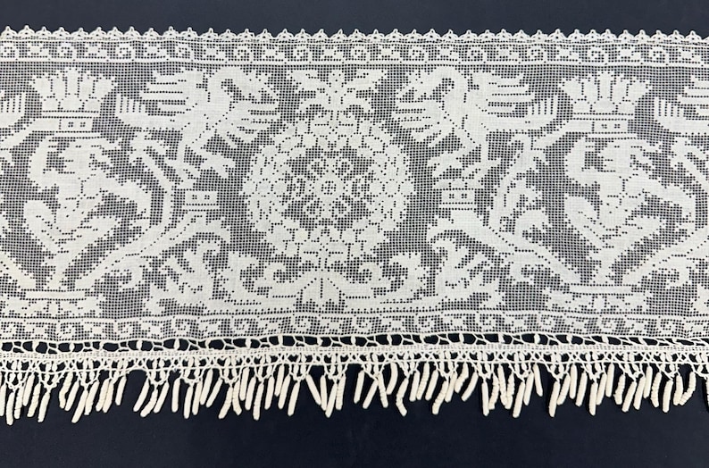 May include: Close-up of a white lace trim with a detailed floral and heraldic design. The lace features a repeating pattern of stylized crowns, floral motifs, and a central medallion. The bottom edge has a fringe.