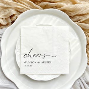 May include: A square white paper napkin featuring the black printed text "cheers" in a cursive script, followed by the names "MADISON & AUSTIN" and the date "10.10.26" in a clean, sans-serif font, centred on the napkin surface.