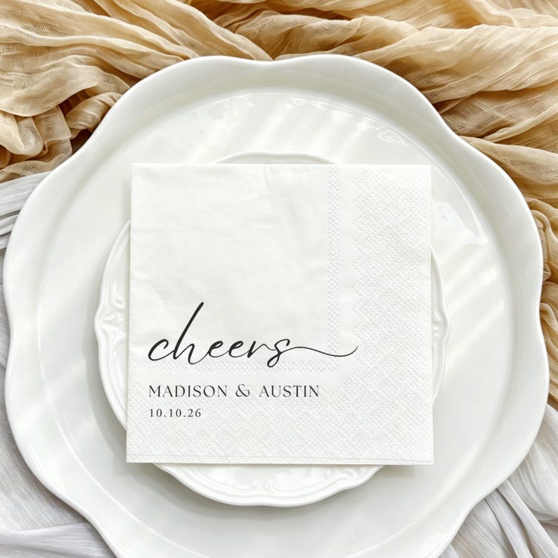 May include: A square white paper napkin featuring the black printed text "cheers" in a cursive script, followed by the names "MADISON & AUSTIN" and the date "10.10.26" in a clean, sans-serif font, centred on the napkin surface.