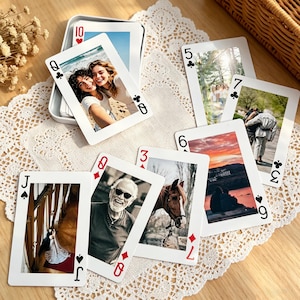 May include: A deck of playing cards with custom photos. The cards are arranged on a white lace doily. The cards are in a silver tin. The photos include people, a horse, and a sunset. The cards are a mix of suits and numbers.