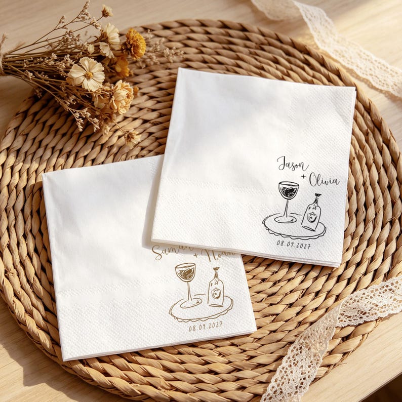 May include: Two square white paper napkins featuring black and gold ink illustrations of a wine glass and a bottle on a tray. The top napkin displays the text Jason + Olivia and the date 08.09.2027, while the bottom napkin shows Saman + Noah.