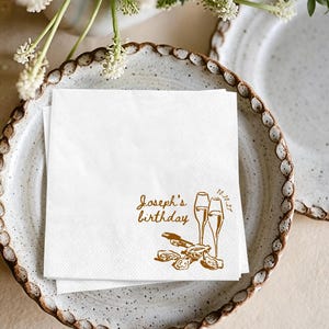 May include: A square white paper napkin featuring gold-coloured ink printing. The design includes the text "Joseph's birthday" and "10.10.27" alongside illustrations of two tall champagne flutes and a cluster of three open oysters with a shucking knife.