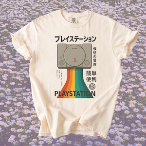 May include: A cream-coloured t-shirt with a PlayStation console graphic and rainbow design. Japanese text is above and below the console, with "PLAYSTATION" at the bottom. The shirt has short sleeves and a crew neck.