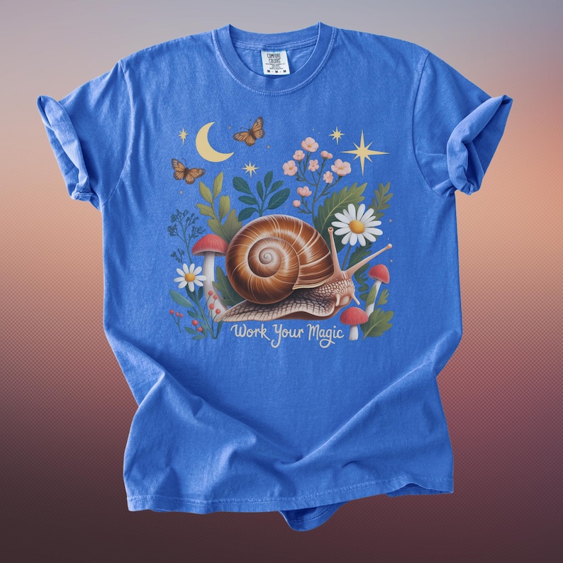 May include: A blue t-shirt featuring a whimsical illustration. The design includes a snail with a brown shell, surrounded by flowers, mushrooms, butterflies, and a crescent moon. The text "Work Your Magic" is below the snail.