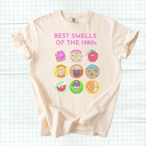 Puede incluir: Camiseta color crema con el texto rosa "BEST SMELLS OF THE 1980s". La camiseta presenta gráficos de dibujos animados coloridos de varios alimentos y golosinas, incluyendo una porción de pizza y un helado.