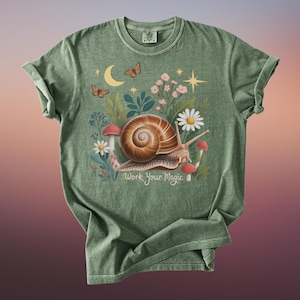 May include: Green t-shirt featuring a whimsical illustration of a snail with a brown shell, surrounded by flowers, mushrooms, and butterflies. The text "Work Your Magic" is below the snail. The shirt has rolled-up sleeves.