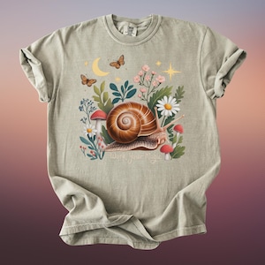 May include: A sage green t-shirt featuring a whimsical illustration of a snail with a brown shell, surrounded by flowers, mushrooms, and butterflies. The text "Work Your Magic" is below the snail.