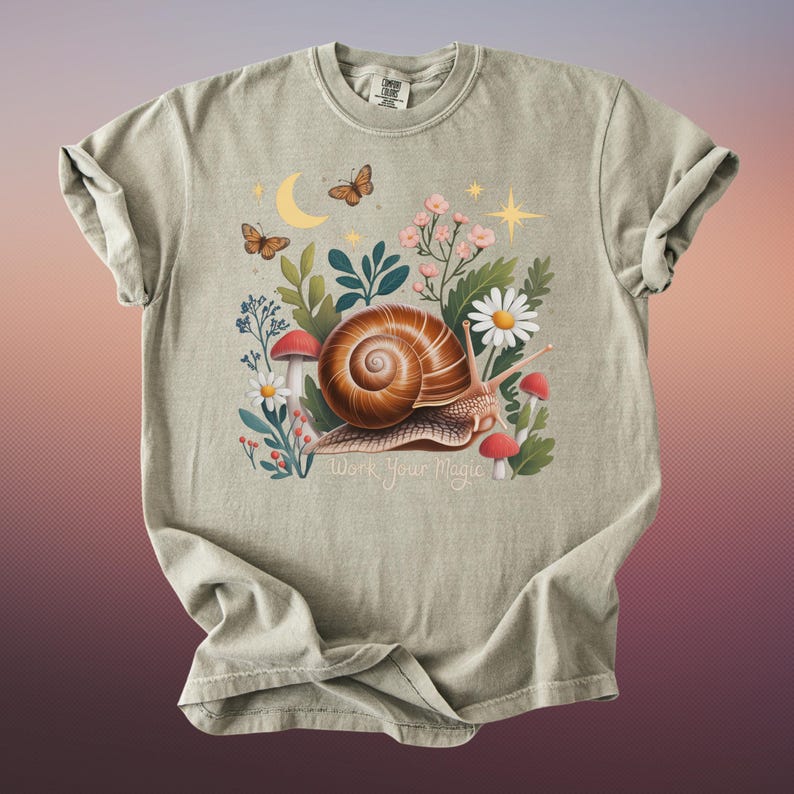 May include: A sage green t-shirt featuring a whimsical illustration of a snail with a brown shell, surrounded by flowers, mushrooms, and butterflies. The text "Work Your Magic" is below the snail.