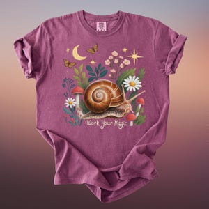 May include: A maroon t-shirt featuring a whimsical illustration of a snail with a spiral shell, surrounded by flowers, mushrooms, and butterflies. The text "Work Your Magic" is below the snail. The shirt has a crew neck and short sleeves.