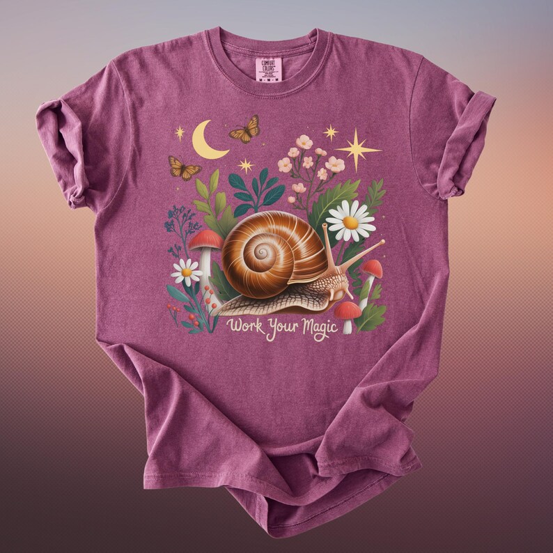 May include: A maroon t-shirt featuring a whimsical illustration of a snail with a spiral shell, surrounded by flowers, mushrooms, and butterflies. The text "Work Your Magic" is below the snail. The shirt has a crew neck and short sleeves.