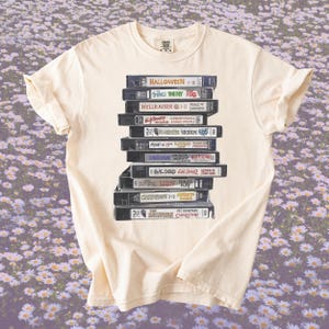 Puede incluir: Camiseta color crema con una pila de cintas VHS con títulos de películas de terror como "Halloween" y "The Fly". La camiseta tiene cuello redondo y mangas cortas, perfecta para un look casual.