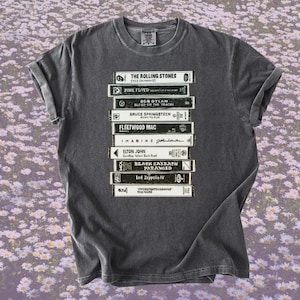 May include: Dark gray t-shirt featuring a stack of vintage cassette tapes with band names like The Rolling Stones, Pink Floyd, and Led Zeppelin. The text on the tapes is in white and black. The shirt is a classic crew neck.