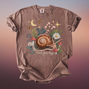 May include: Brown t-shirt featuring a whimsical illustration of a snail with a spiral shell, surrounded by flowers, mushrooms, and butterflies. The text "Work Your Magic" is below the snail. The shirt has a crew neck and rolled sleeves.