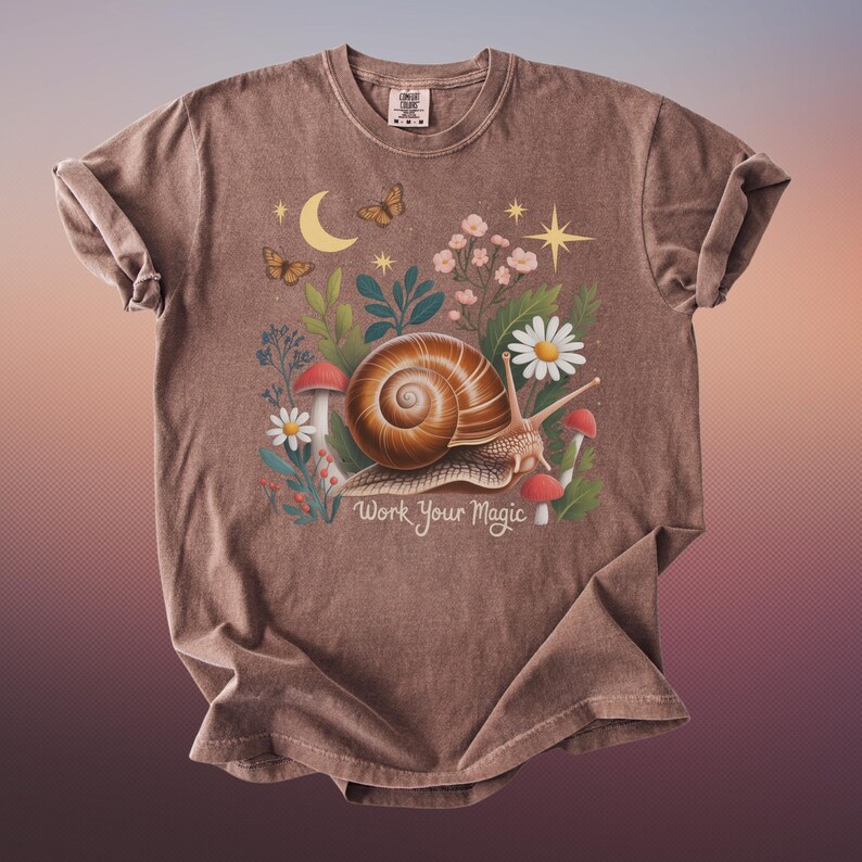 May include: Brown t-shirt featuring a whimsical illustration of a snail with a spiral shell, surrounded by flowers, mushrooms, and butterflies. The text "Work Your Magic" is below the snail. The shirt has a crew neck and rolled sleeves.