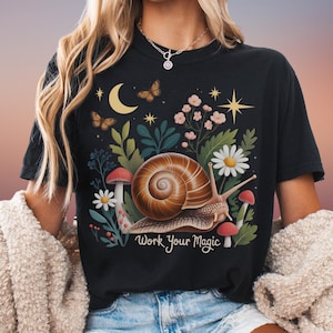 May include: Black t-shirt featuring a whimsical design with a snail, mushrooms, flowers, butterflies, and a crescent moon. The text "Work Your Magic" is printed below the snail. The design is in shades of brown, green, pink, and yellow.