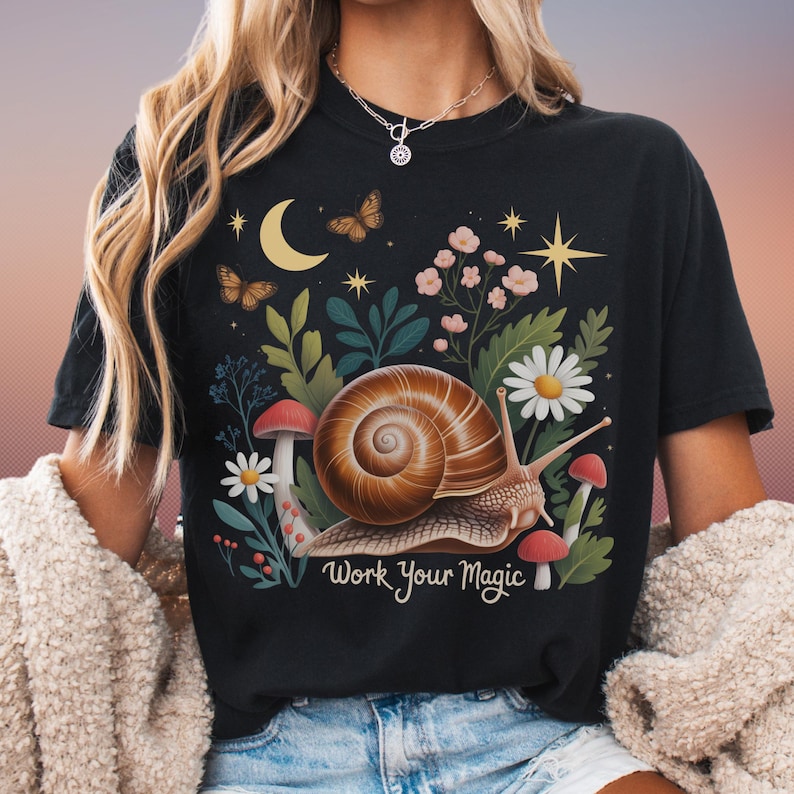 May include: Black t-shirt featuring a whimsical design with a snail, mushrooms, flowers, butterflies, and a crescent moon. The text "Work Your Magic" is printed below the snail. The design is in shades of brown, green, pink, and yellow.
