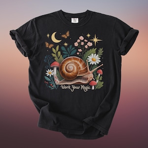 May include: Black t-shirt featuring a whimsical illustration of a snail with a brown shell, surrounded by flowers, mushrooms, and butterflies. The text "Work Your Magic" is below the snail. The shirt has rolled-up sleeves.