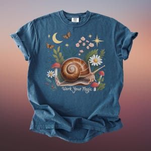 May include: A blue t-shirt featuring a whimsical illustration of a snail with a brown shell, surrounded by flowers, mushrooms, butterflies, and a crescent moon. The text "Work Your Magic" is below the snail.