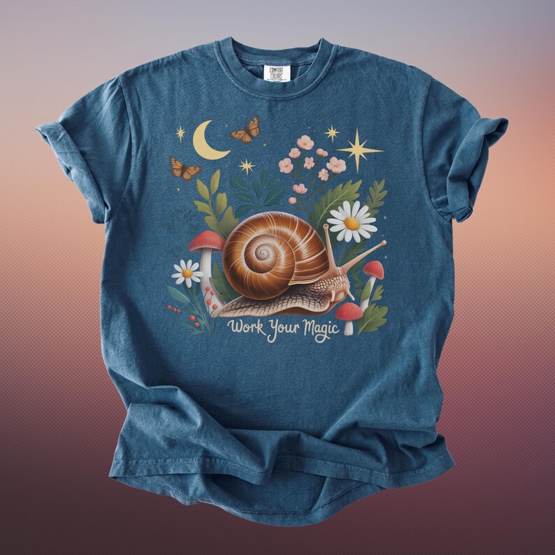 May include: A blue t-shirt featuring a whimsical illustration of a snail with a brown shell, surrounded by flowers, mushrooms, butterflies, and a crescent moon. The text "Work Your Magic" is below the snail.