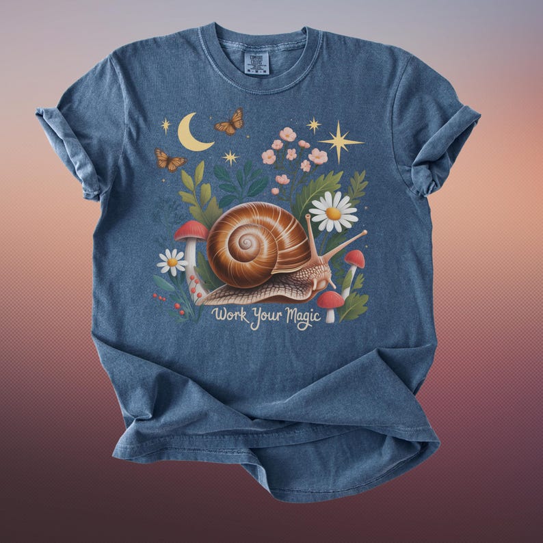 May include: A blue t-shirt featuring a whimsical illustration of a snail with a brown shell, surrounded by flowers, mushrooms, and butterflies. The text "Work Your Magic" is printed below the snail. The shirt has rolled-up sleeves.
